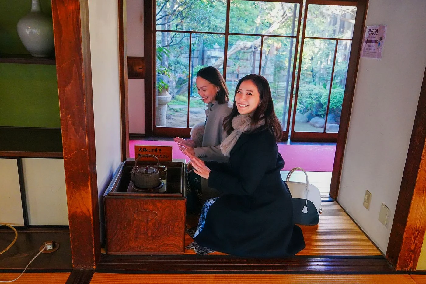 Two visitors warming their hands over a hibachi brazier at Yamamoto-tei, with the garden outside